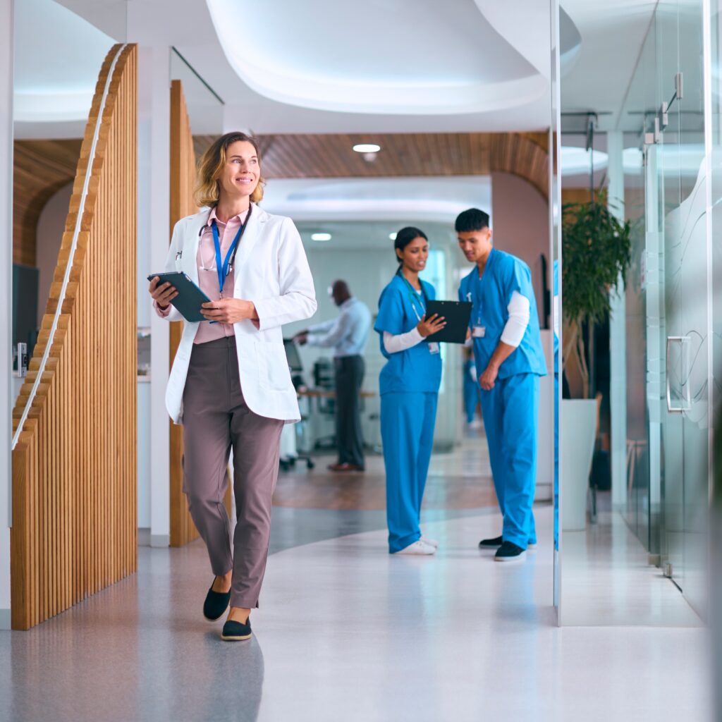 Female Doctor In White Coat With Digital Tablet In Hospital With Colleagues In Background